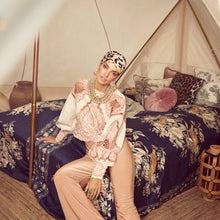 Load image into Gallery viewer, A woman models a bohemian-style outfit consisting of a light pink embroidered blouse, beige trousers, and a patterned headscarf while sitting on a bed inside a canvas tent.