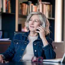Load image into Gallery viewer, Woman wearing a wig for hair loss. "Liz" by La Vida Wigs, talking on a phone in an office setting with books and a laptop.