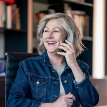 Load image into Gallery viewer, Woman wearing a wig for hair loss. "Liz" by La Vida Wigs, talking on a phone in an office setting with books and a laptop.