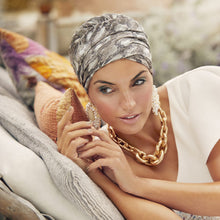 Load image into Gallery viewer, A woman reclining on a cushioned surface, wearing a patterned gray and white Sapphire Boho Turban, a white sleeveless dress, and a chunky gold chain necklace.